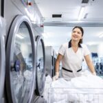 Female dry cleaning worker pushing laundry cart by washing machine.