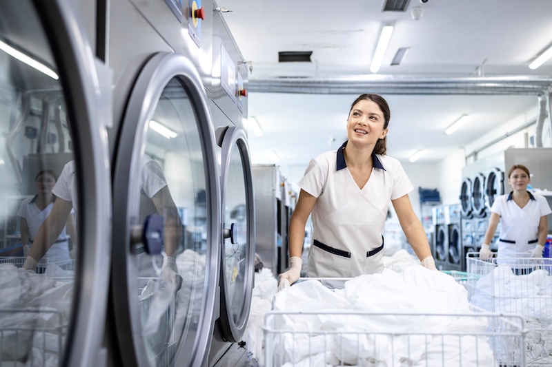 Female dry cleaning worker pushing laundry cart by washing machine.