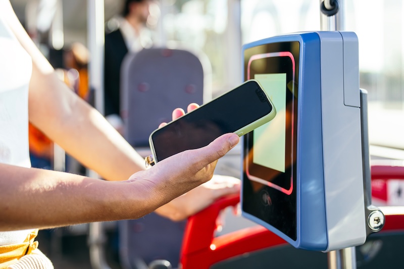 NFC Forum Woman using mobile payment on a contactless reader inside a public city bus, representing modern travel and technology
