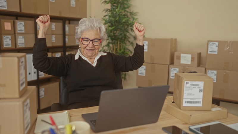 Reelables Senior woman raises fists at laptop amid stacked shipping boxes and labels on shelves in a building; entrepreneurial pride.